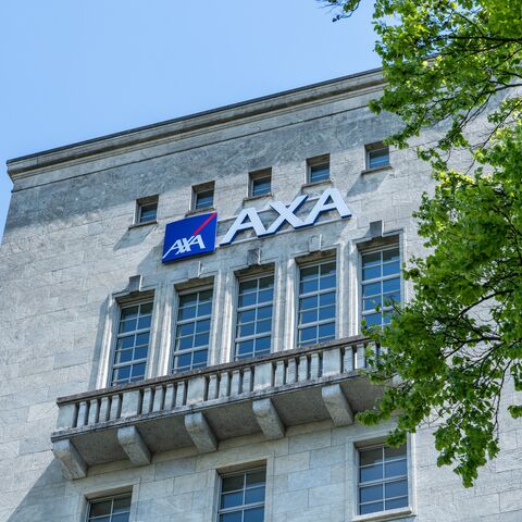 La photo montre la façade d’un bâtiment avec le logo AXA et plusieurs fenêtres, à côté d’un grand arbre aux feuilles vertes.