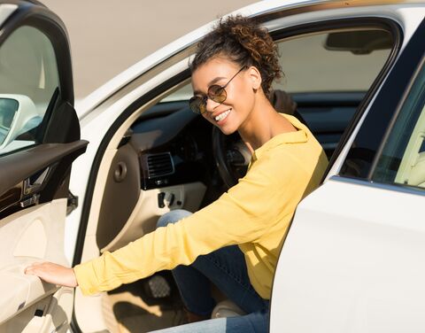 Eine junge Frau mit Sonnenbrille und gelber Jacke steigt lachend aus ihrem weissen Auto.