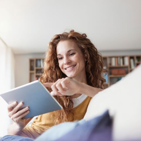 Una donna sorridente con i capelli ricci è seduta su un divano e utilizza un tablet, circondata da libri in una stanza luminosa. 
