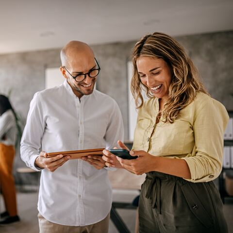 Two people in a modern office are smiling while looking at a smartphone together; colleagues are working in the background.