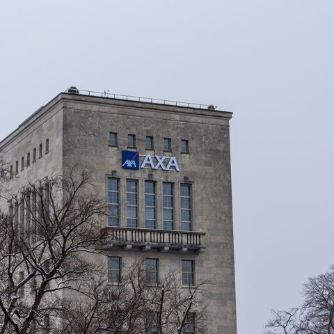 Un edificio grigio con il logo AXA sopra diverse grandi finestre, circondato da alberi spogli e sotto un cielo nuvoloso.