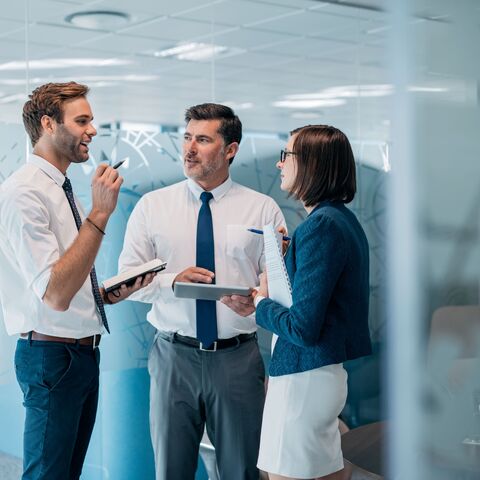 Tre uomini d'affari in abito formale sono in piedi in un ufficio moderno, discutono e tengono in mano taccuini e tablet.