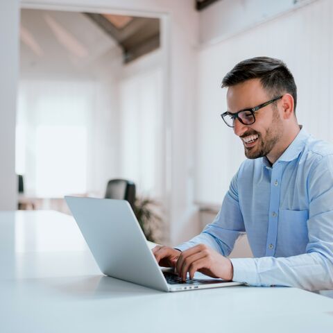 Ein lächelnder Mann mit Brille und blauem Hemd arbeitet an einem Laptop in einem hellen, modernen Büro mit grossen Fenstern.