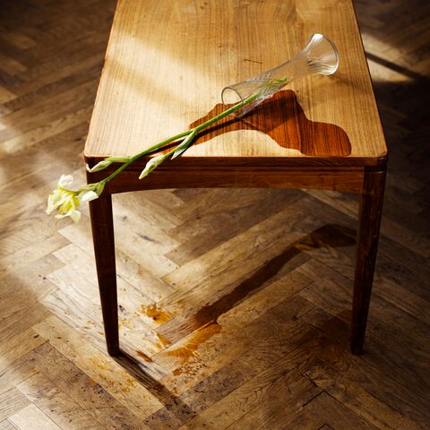 Auf einem kleinen Tisch steht eine umgekippte Blumenvase. Das Wasser tropft vom Tisch und bildet auf dem Parkett eine Pfütze.