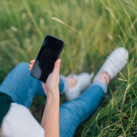Eine Person mit Kopfhörern sitzt auf einer Wiese, hält ein Smartphone in der Hand und trägt Jeans sowie weisse Sneakers.
