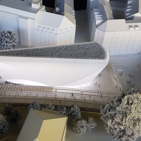 Ein architektonisches Modell zeigt ein modernes Gebäude mit spitzem Dach inmitten eines Stadtteils mit Bäumen und umliegenden Gebäuden.