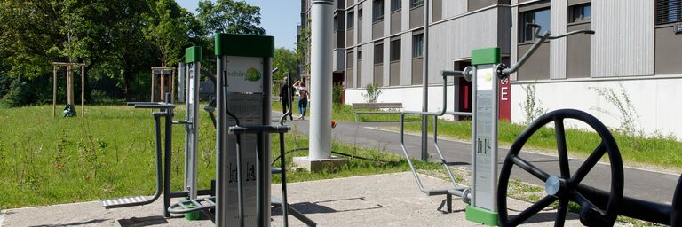 Area fitness all'aperto con varie attrezzature per l'allenamento, circondata da aree verdi e da un edificio moderno sullo sfondo.