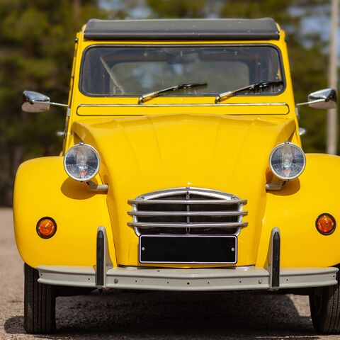 Un'auto d'epoca di colore giallo brillante con fari tondi e targa nera si trova in un parcheggio su uno sfondo sfocato.