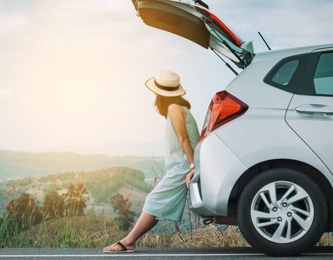 Eine Frau mit Hut lehnt sich an den geöffneten Kofferraum eines silbernen Autos und blickt auf eine hügelige Landschaft im Sonnenuntergang.