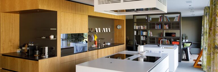 Cucina moderna con armadi in legno, piani di lavoro bianchi e un mobile centrale, con una libreria e un tavolo da pranzo sullo sfondo.