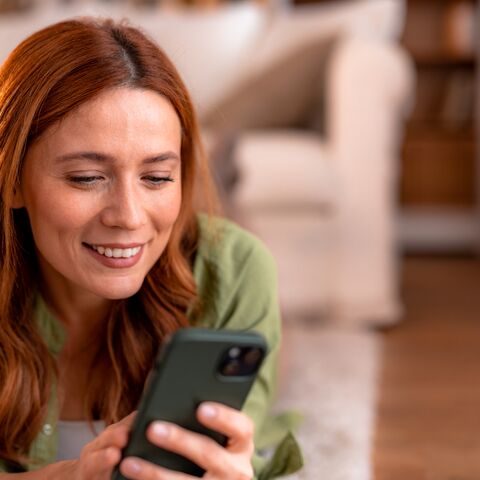Woman is lying on the floor looking at her cell phone with a smile. A white sofa can be seen in the background.