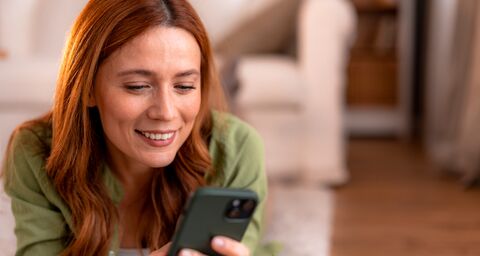 Une femme est allongée par terre et regarde son téléphone portable en souriant. À l’arrière-plan, on voit un canapé blanc.