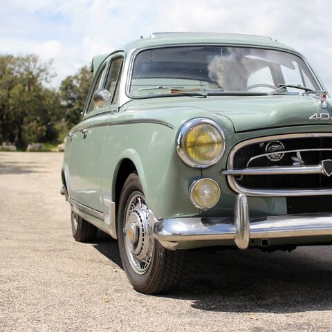Un'auto d'epoca verde chiaro è parcheggiata su una strada in pieno sole, circondata da alberi e da un cielo leggermente nuvoloso.
