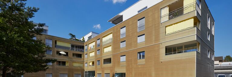 Immeuble locatif moderne avec façade en bois, volets roulants jaunes et balcons par une journée ensoleillée avec ciel bleu.