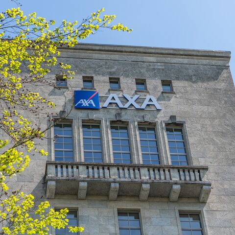 Ansicht eines grossen, grauen Gebäudes mit dem AXA-Logo, umgeben von grünen, blühenden Bäumen unter einem klaren blauen Himmel.