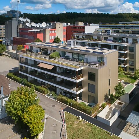 Edificio residenziale a più piani con balconi verdi e facciate moderne, circondato da altri edifici e spazi verdi sotto un cielo azzurro.