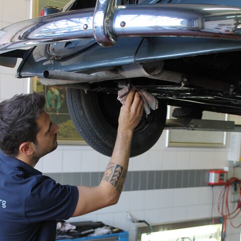 Un meccanico con un braccio tatuato pulisce sotto un'auto sollevata in un'officina con un armadio degli attrezzi e pareti sullo sfondo.