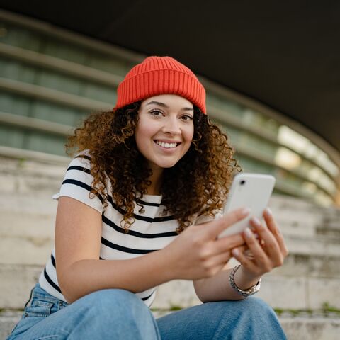 Eine junge Frau steht in ihrem Wohnzimmer und schaut auf ihr Handy. Im Hintergrund sind ein Bücherregal und ein Esstisch zu sehen.