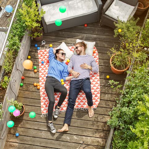 A young couple are relaxing on their balcony, lying on a blanket and holding hands.