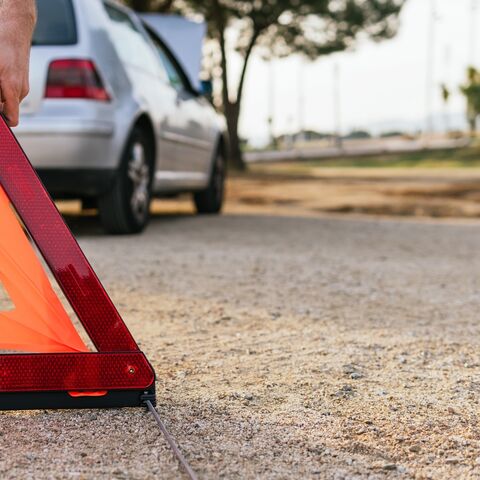 Su una strada, davanti a un’auto parcheggiata è collocato un triangolo di emergenza rosso, mentre accanto si intravede una persona.