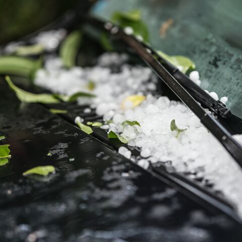 Gros plan d’un essuie-glace de voiture avec de la grêle et des feuilles vertes sur le pare-brise mouillé après une tempête de grêle.
