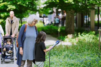 A child and an older woman look with interest at an information board. In the background, a man pushes a baby carriage.