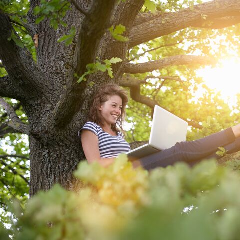 Una giovane donna siede sorridente su un albero e lavora su un laptop alla luce del sole, circondata da foglie verdi.