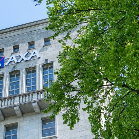 Das Bild zeigt die Fassade eines Gebäudes mit dem AXA-Logo und mehreren Fenstern, daneben ein grosser Baum mit grünen Blättern.