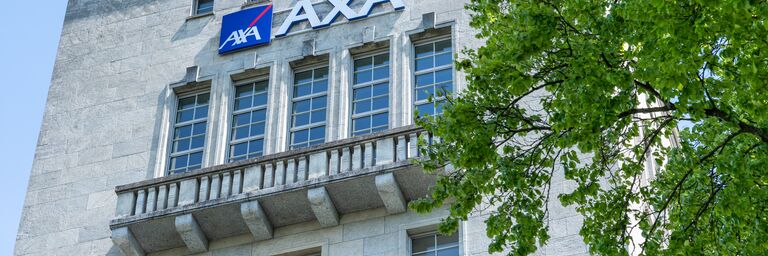 L’immagine mostra la facciata di un edificio con il logo AXA e diverse finestre, accanto a un grande albero con foglie verdi.