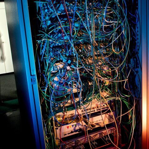 Ein Mann steht in einem dunklen Raum vor einem Server-Rack, das mit zahlreichen Kabeln und leuchtenden Komponenten überfüllt ist.