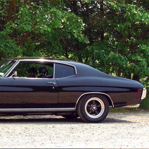 Una coupé d'epoca nera e lucida è parcheggiata su una pista di ghiaia davanti ad alberi verdeggianti, con cerchi lucidi e un design classico.