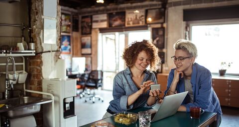  Deux femmes souriantes dans un bureau confortable regardent ensemble un smartphone alors qu'elles sont assises à une table avec un ordinateur portable et des snacks.