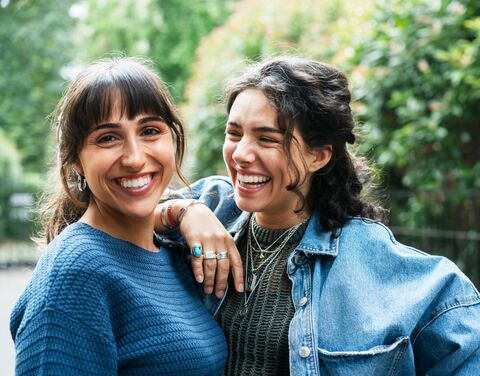 Two young women are standing in a sunny park, laughing heartily together.
