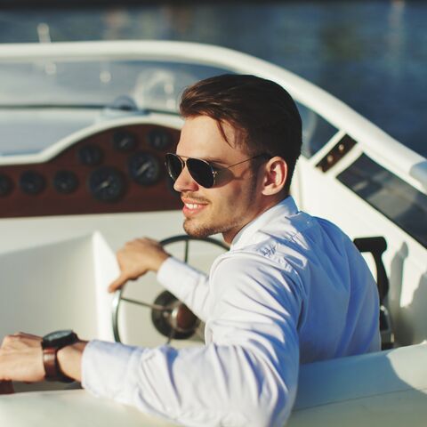A man with sunglasses and a white shirt steers his motorboat over the lake in the evening sun.
