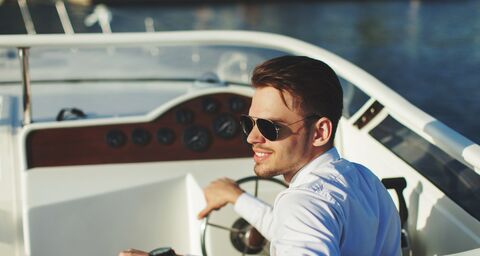 A man with sunglasses and a white shirt steers his motorboat over the lake in the evening sun.