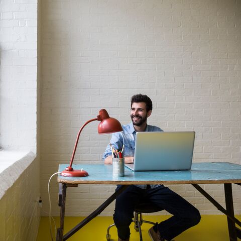 Ein Mann sitzt lächelnd an einem Schreibtisch mit einem Laptop, einer roten Lampe und Stiften, neben einem grossen Fenster in einem hellen Raum.