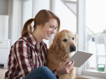 Une jeune femme souriante est assise par terre et regarde une tablette avec son chien