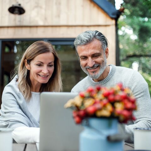 A smiling couple is sitting outside at a table in front of a laptop, with a vase full of colorful flowers in the foreground.
