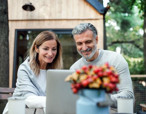 Ein lächelndes Paar sitzt draussen an einem Tisch vor einem Laptop, mit einer Vase voller bunter Blumen im Vordergrund.
