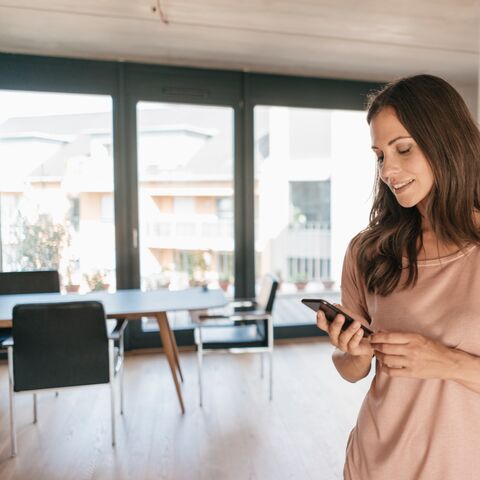 Eine junge Frau steht in ihrem Wohnzimmer und schaut auf ihr Handy. Im Hintergrund sind ein Bücherregal und ein Esstisch zu sehen.