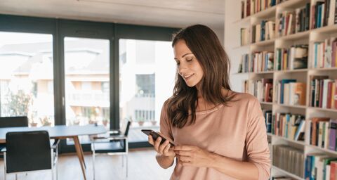 Una giovane donna è in salotto e guarda il suo cellulare. Sullo sfondo si vedono una libreria e un tavolo da pranzo.