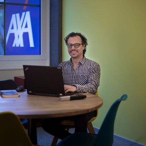 Ein Mann mit Brille sitzt lächelnd an einem runden Tisch vor einem Laptop in einem Büro mit einem AXA-Logo im Fenster im Hintergrund.