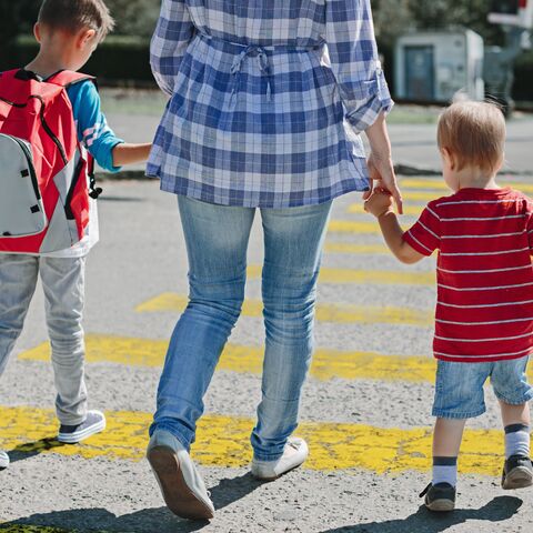 Eine erwachsene Person führt zwei Kinder, eines mit rotem Rucksack, über einen Zebrastreifen in einer städtischen Umgebung.