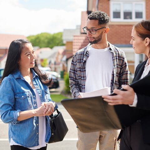 Une femme en costume montre des documents à un couple en tenue décontractée, dehors, devant un immeuble d'habitation, tandis que tout le monde se parle en souriant.
