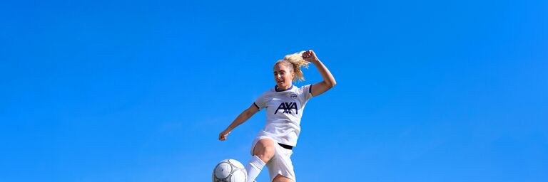 Eine blonde Fussballspielerin im weissen AXA-Trikot springt vor strahlend blauem Himmel hoch in die Luft und kontrolliert den Ball mit dem Fuss.