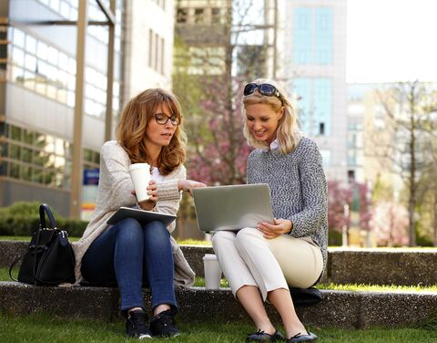Due donne sono sedute all’aperto su un gradino in pietra, bevono caffè e lavorano insieme a un laptop, circondate da edifici urbani.
