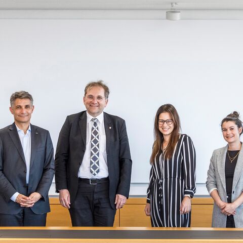Sechs lächelnde Personen in formeller Kleidung stehen nebeneinander vor einer weissen Wand in einem Konferenzraum.