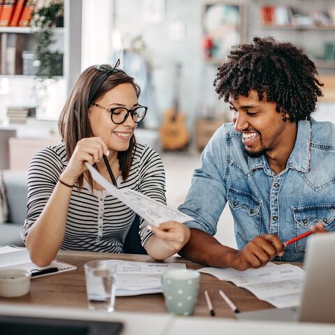Un couple souriant est assis à une table, regarde des documents et travaille ensemble; en arrière-plan, on voit des étagères et des décorations.