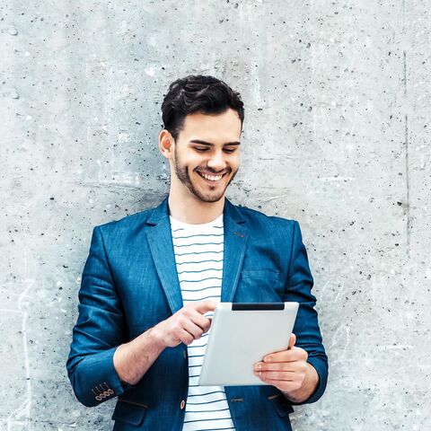Un uomo in blazer blu e maglietta a righe sorride mentre usa un tablet davanti a un muro di cemento grigio.