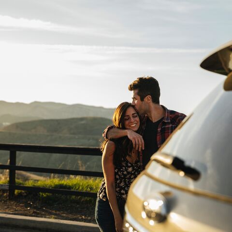 Un couple s’embrasse amoureusement à côté d’une voiture, avec un paysage montagneux pittoresque et un coucher de soleil en arrière-plan.